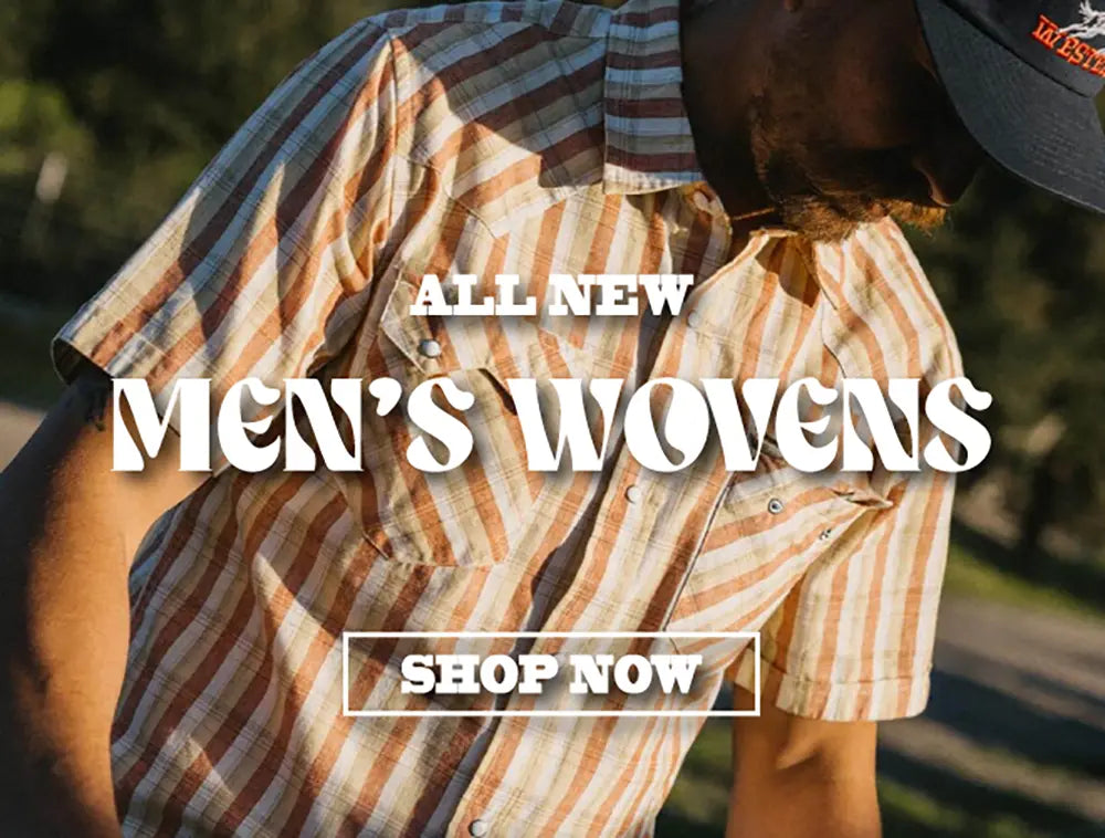 A man wearing a short-sleeve plaid button-up shirt in warm tan, brown, and cream tones, with large overlaid text reading 'ALL NEW MEN'S WOVENS' and a 'SHOP NOW' button on a blurred outdoor background.
