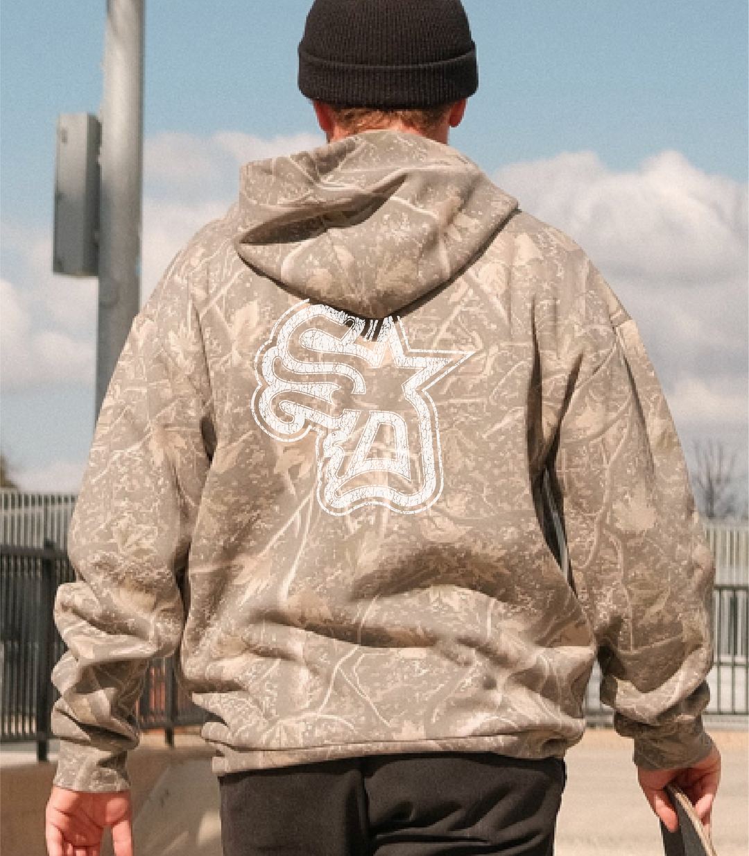 Person wearing a camouflage hoodie with a logo on the back, standing outdoors.