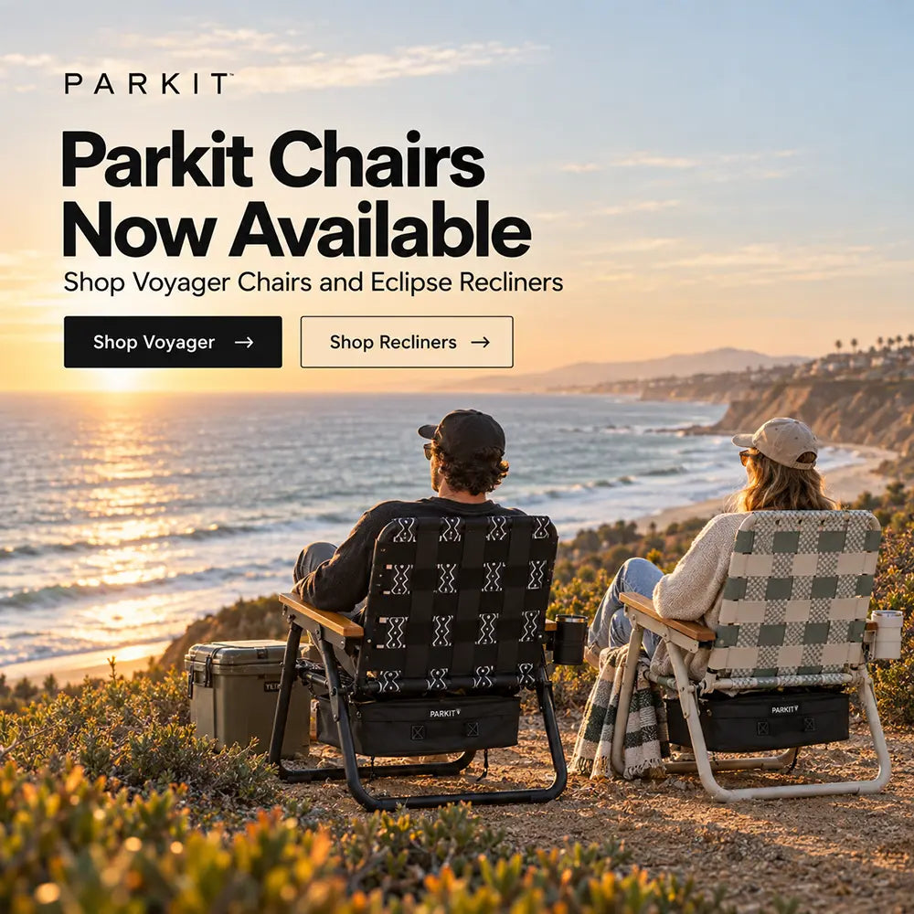 Two people sitting in Parkit outdoor chairs on a coastal cliffside overlooking the ocean at sunset. Large promotional text on the left reads 'PARKIT Parkit Chairs Now Available' with buttons to shop Voyager chairs and Eclipse recliners.