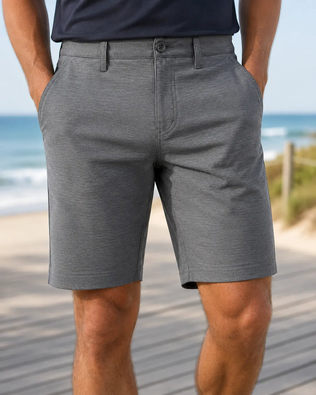A man wearing heather gray athletic shorts and a dark navy t-shirt, standing on a wooden boardwalk with a beach and ocean in the background.