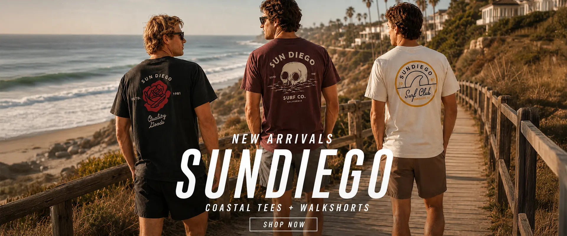 Three men wearing graphic tees and shorts walk along a wooden boardwalk overlooking the ocean. The tees feature the brand name 'Sundiego' and coastal graphics, with text overlay reading 'NEW ARRIVALS SUNDIEGO COASTAL TEES + WALKSHOOTS'.