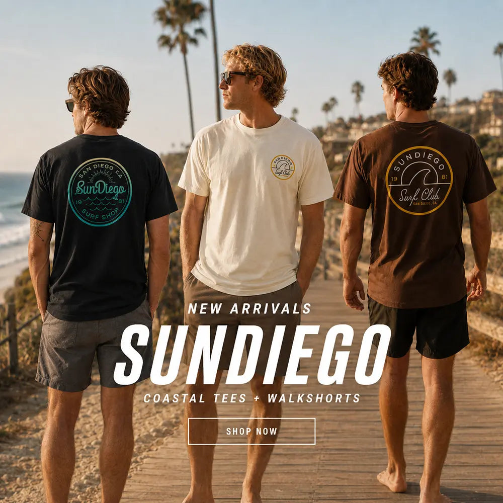 Three men standing on a wooden boardwalk by the beach, shown from the back to display Sundiego t-shirts and walkshorts. The left man wears a black t-shirt with a teal circular 'Sun Diego' graphic, the center man wears a cream t-shirt with a small yellow graphic, and the right man wears a brown t-shirt with a yellow circular 'Sun Diego Surf Club' graphic.