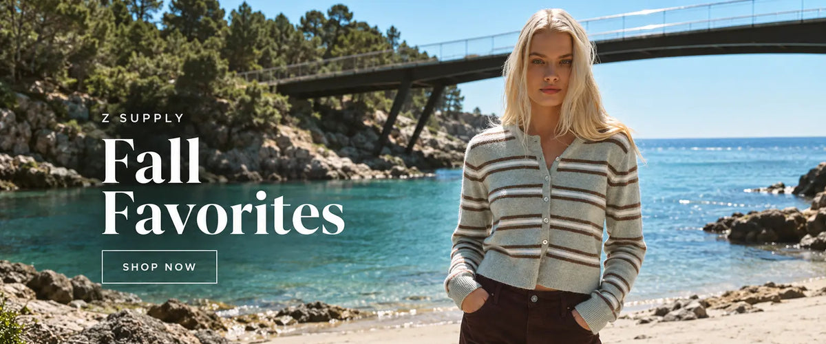 A woman stands on a rocky beach with turquoise water and a bridge in the background, wearing a cream cardigan with brown stripes. Large text on the left reads '2 SUPPLY Fall Favorites' with a 'SHOP NOW' button. 